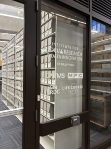 Looking in the window of the Institute for Social Research and Data Innovation 50 Willey Hall at the IPUMS archives shelves filled with accordion folders and boxes